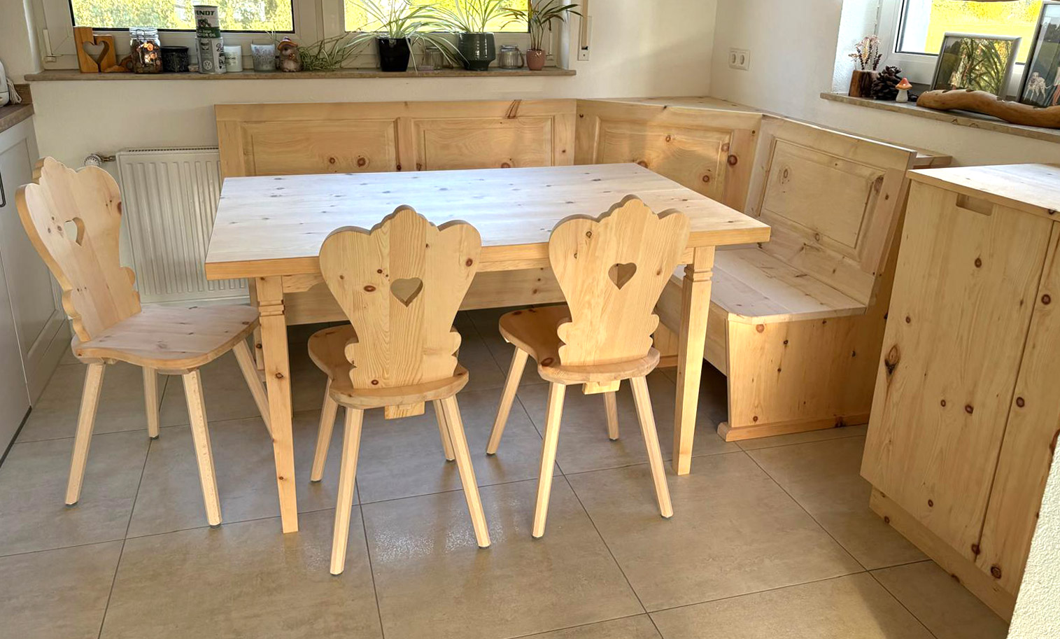 Sitzgruppe aus Zirbe mit Eck-Sitzbank, Tisch und Stühlen mit Herzausschnitt an der Lehne. Seitlich eine Kommode aus Zirbeholz.